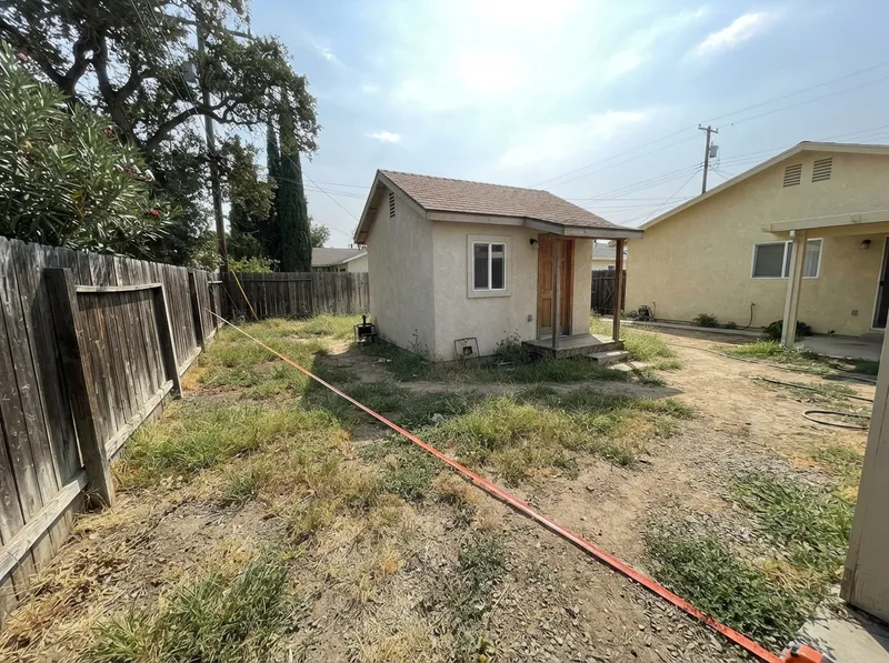 Small detached backyard cottage behind a stucco house, with a surveyor's tape measure stretched across the yard toward the side fence