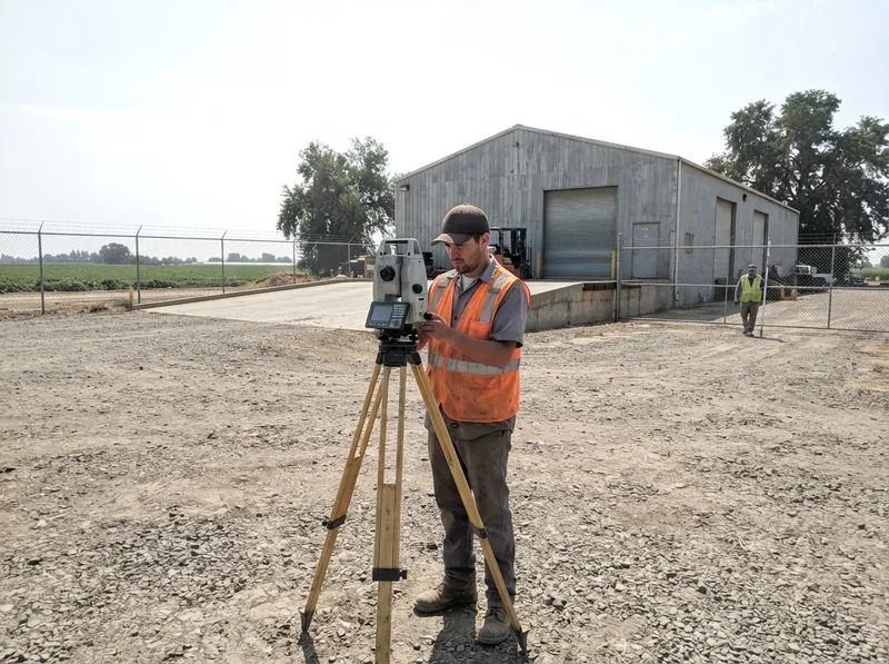Land surveyor operating a total station on the gravel yard of a commercial agricultural warehouse, with a chain-link fence and farm fields behind
