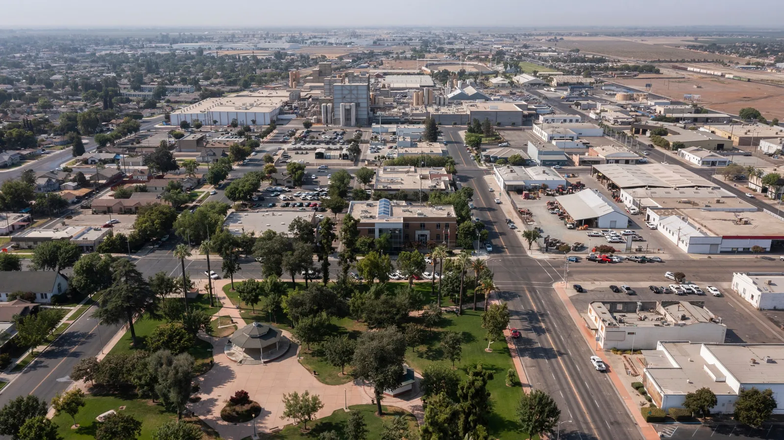 Aerial view of downtown Tulare, California, with a tree-filled park and bandstand in the foreground, civic and commercial buildings, and farmland on the horizon