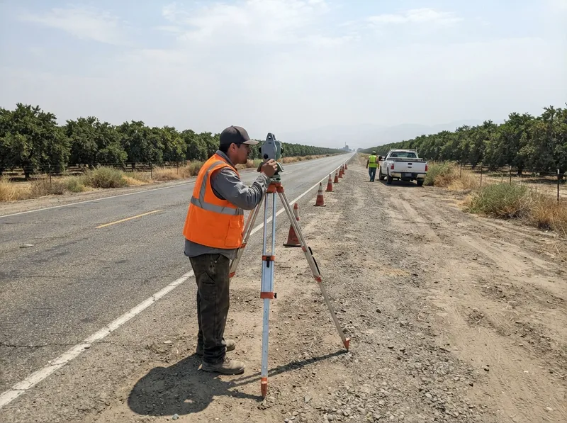 Land surveyor at a total station on the shoulder of a two-lane rural highway between citrus orchards, with traffic cones and a second crew member by a pickup behind